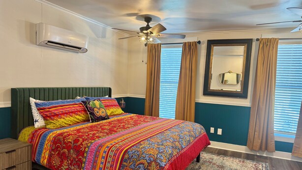 A bedroom with a bed, a ceiling fan, an air conditioning unit, a nightstand, a dresser, and a window with curtains. - Main Street House with private pool! (Bay St. Louis)