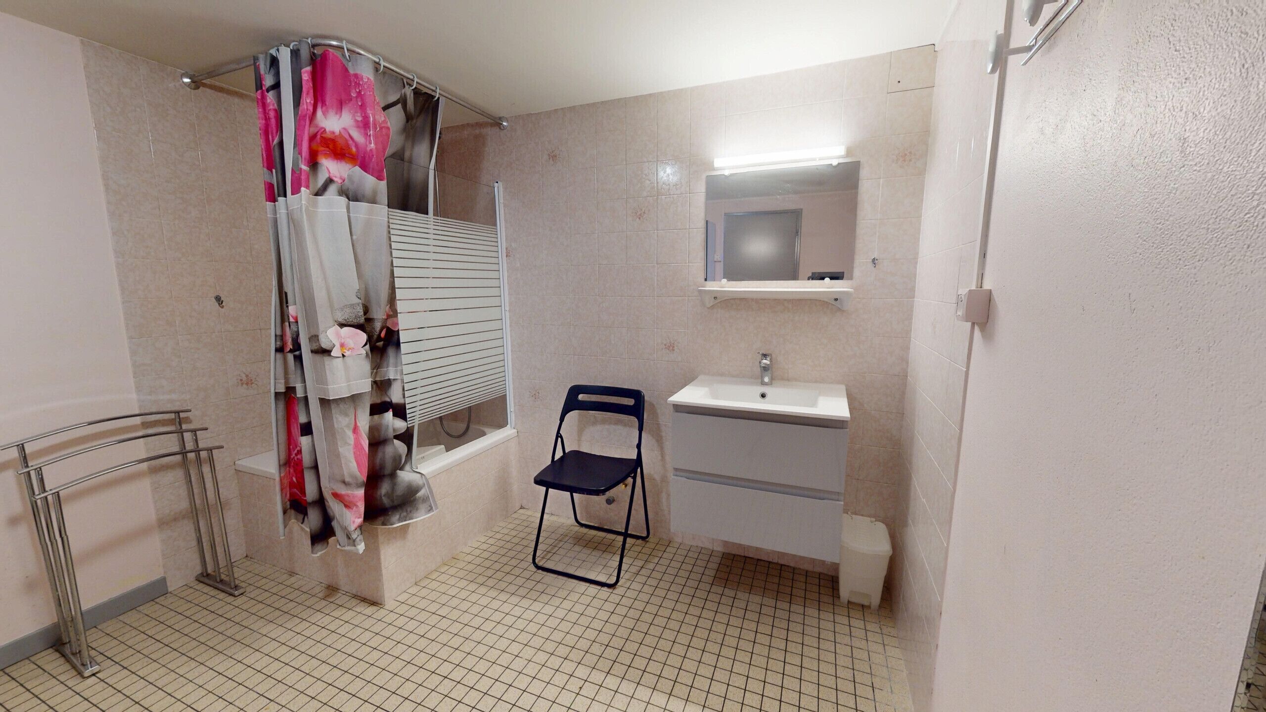 A bathroom with a shower curtain, a sink, a mirror, a chair, and a toilet.