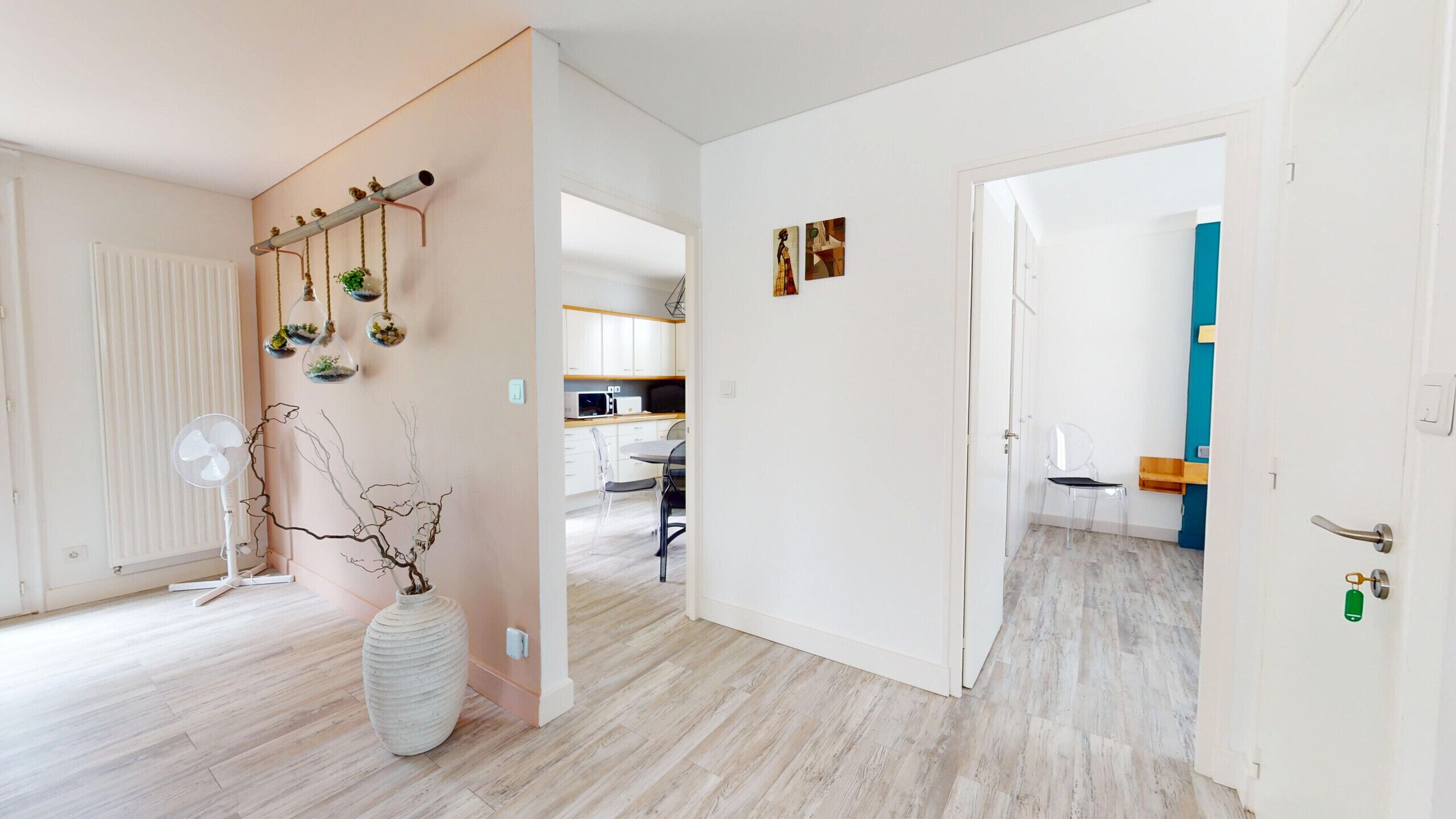 A modern interior with a wooden floor, a white radiator, a large vase with branches, and a kitchen area visible through an open door.