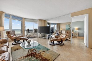 A spacious living room with leather sofas, a glass coffee table, a flat-screen TV, and large windows offering ocean views. - VAN LEE BEACH CONDO 204!!!! (Ormond Beach)