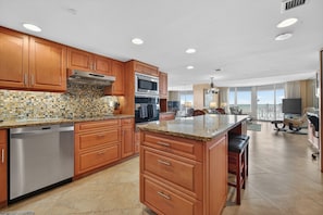 A modern kitchen with wooden cabinets, a granite countertop, stainless steel appliances, and a large island with bar stools. - VAN LEE BEACH CONDO 204!!!! (Ormond Beach)