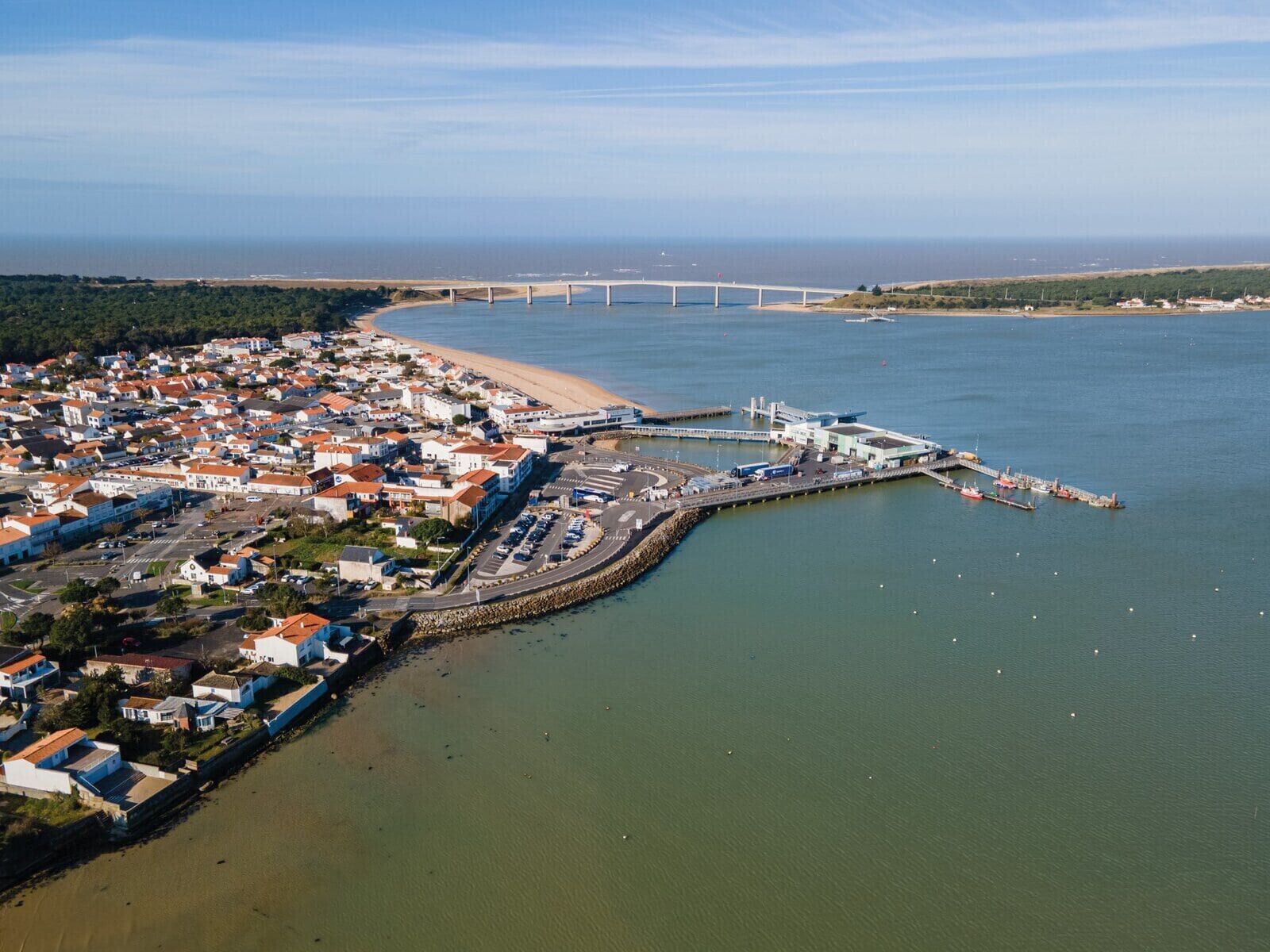 An aerial view of a coastal town with a marina, a bridge in the background, and a curved road leading to the beach.