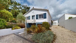 Une maison à deux étages, à l’extérieur blanc et aux volets bleus, entourée de gravier et de verdure.