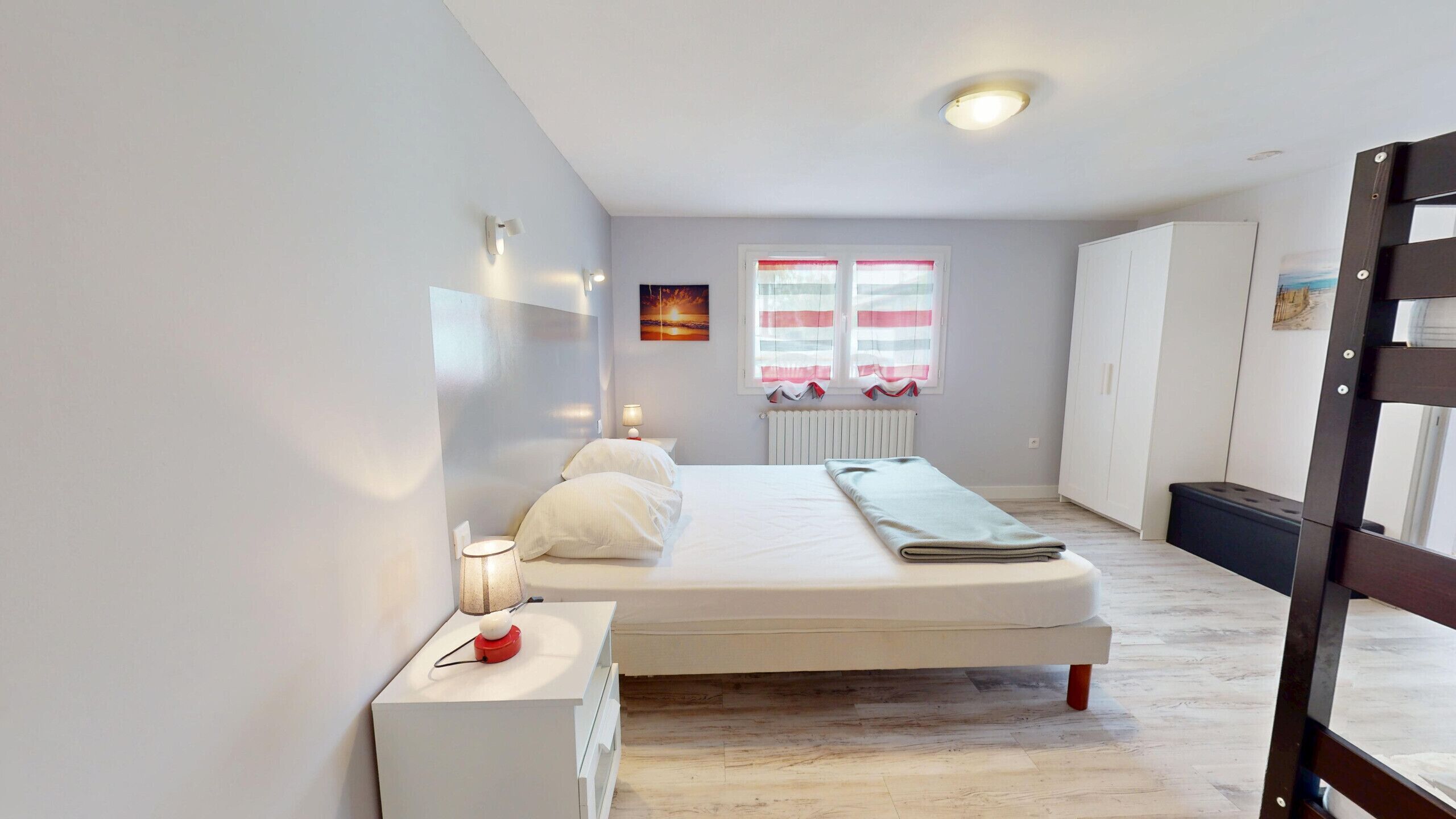 Una camera da letto con un letto, comodino, lampada, finestra con tende, termosifone e vista esterna.