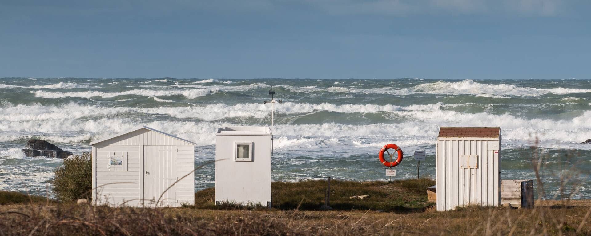 Three small white sheds by the sea with rough waves in the background.