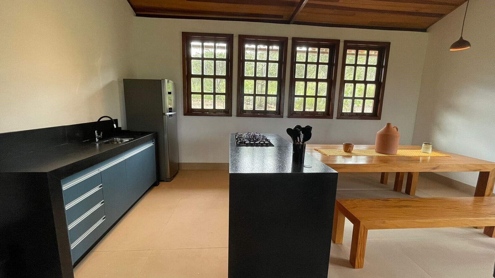 Una cocina moderna con encimera negra, un refrigerador, una mesa de comedor y tres ventanas con vista a follaje.