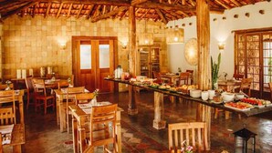Uma área de refeições rústica com mesas e cadeiras de madeira, uma longa mesa comunitária com comida e um alto teto de madeira.