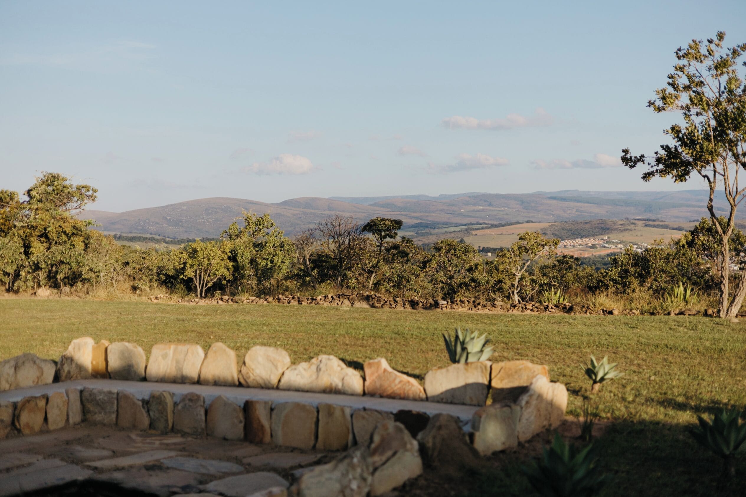 A stone retaining wall with a grassy field and trees in the foreground, and a distant view of hills and a small settlement.