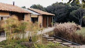 Uma casa rústica com telhado de cerâmica, um lago e vegetação ao redor.