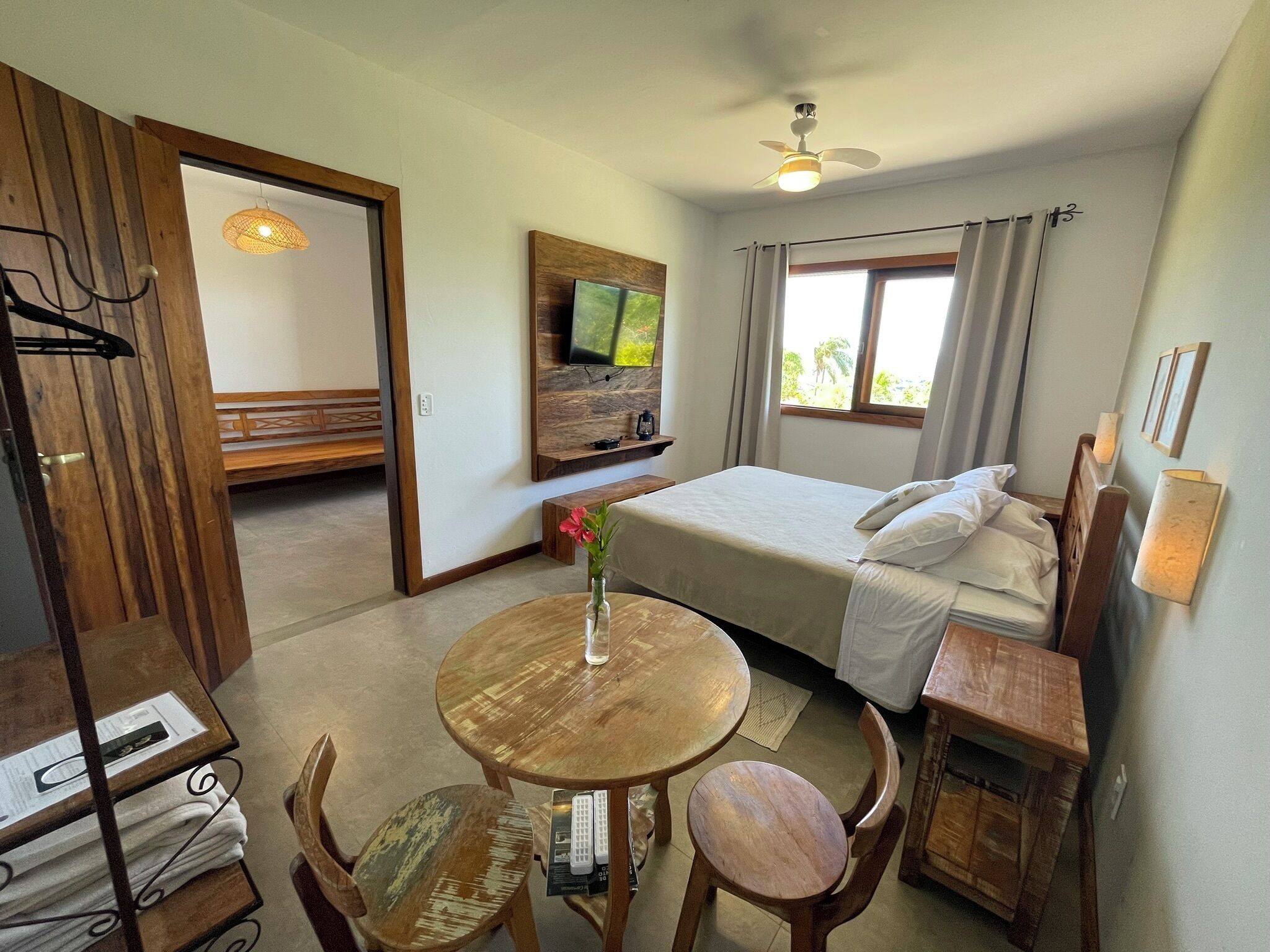 Quarto de hotel com cama, televisão, mesa de jantar com cadeiras, ventilador de teto e janela com cortinas.