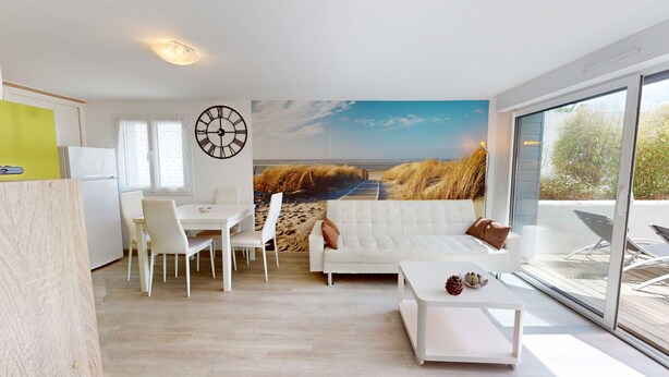 A modern living room with a large window, a white sofa, a dining table with chairs, and a wall clock. - Dune - Apartment 40m² with Terrace (La Barre-de-Monts)