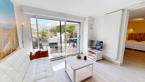 A modern living room with a sofa, a coffee table, a TV, and a sliding glass door leading to an outdoor patio. - Dune - Apartment 40m² with Terrace (La Barre-de-Monts)