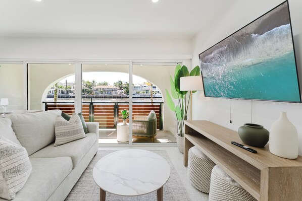 Ein modernes Wohnzimmer mit großem Fernseher, einer Couch, einem Couchtisch und einem Balkon mit Blick auf einen Wasserweg und Gebäude.