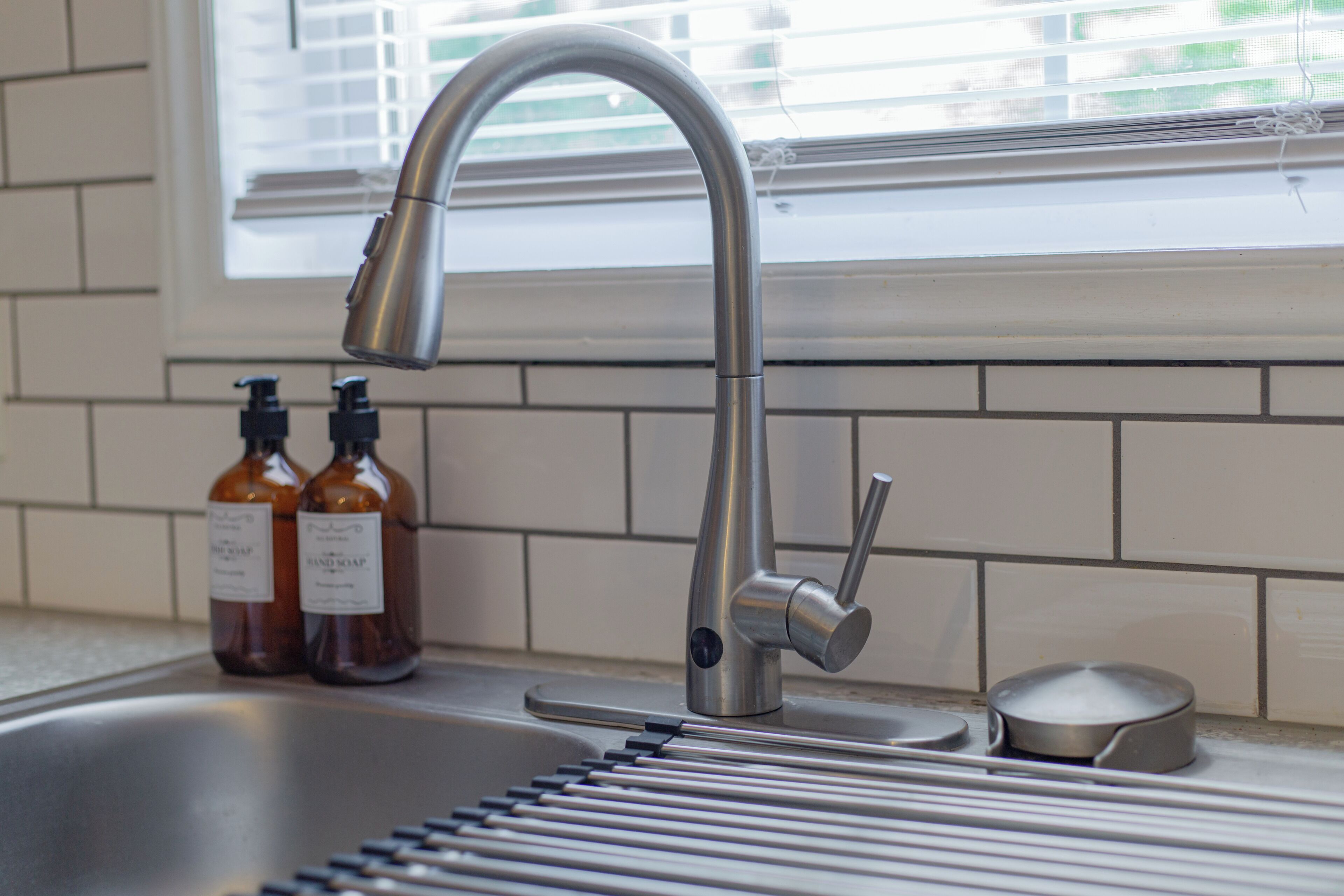A kitchen sink with a modern faucet, two soap bottles, and a window with blinds.