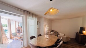 A dining area with a wooden table and chairs, a hanging light fixture, and a view of the outdoors through large windows. - Le cocon de Sète - patio+pool and beach on foot (Sète)