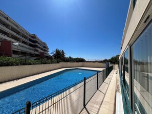 A swimming pool with a metal fence, a building with multiple balconies, and a clear blue sky. - Le cocon de Sète - patio+pool and beach on foot (Sète)