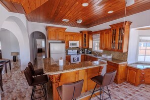 A spacious kitchen with wooden cabinets, granite countertops, and a central island with bar stools. - Golden Pelican – Ocean Front Lower Level Suite on North Caicos (North Caicos)