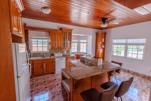 Cucina con mobili in legno, piani in granito, isola centrale con sedute, ventilatore a soffitto e finestre con vista.