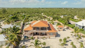 Une grande maison dotée d’un balcon, entourée de palmiers et bordée d’une zone sablonneuse.