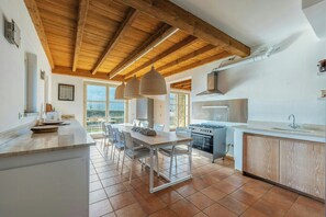 Une cuisine avec un plafond en bois, un coin repas avec des chaises, une grande fenêtre et un poêle.