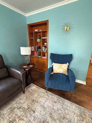 Ein Wohnzimmer mit einem dunkelbraunen Ledersofa, einem blauen Sessel mit floralem Kissen, einem Holzregal und einer Wanduhr.