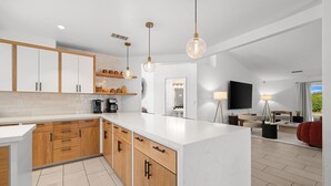 A modern kitchen with wooden cabinets, a marble island, pendant lighting, and a dining area with a TV and seating. - Sandpiper by Nest Villas | 5BR Estate | Large Pool (Indio)