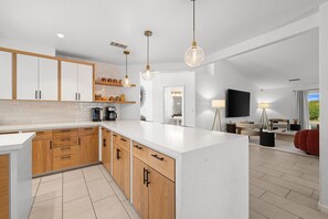 A modern kitchen with wooden cabinets, a marble island, pendant lighting, and a dining area with a TV and seating. - Sandpiper by Nest Villas | 5BR Estate | Large Pool (Indio)