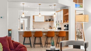 A modern kitchen with bar stools, pendant lighting, and a dining area with a red sofa and a grey chair. - Sandpiper by Nest Villas | 5BR Estate | Large Pool (Indio)