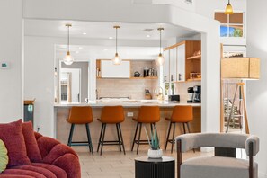 A modern kitchen with bar stools, pendant lighting, and a dining area with a red sofa and a grey chair. - Sandpiper by Nest Villas | 5BR Estate | Large Pool (Indio)