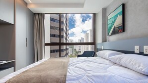 A hotel room with a bed, a window with a view of buildings, a chair, and a painting on the wall. - Rooftop#1101 - Luxury Apartment By Carpediem (Recife)