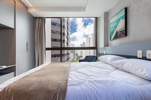 Ein Hotelzimmer mit Bett, Fenster mit Blick auf Gebäude, einem Sessel und einem Bild an der Wand.
