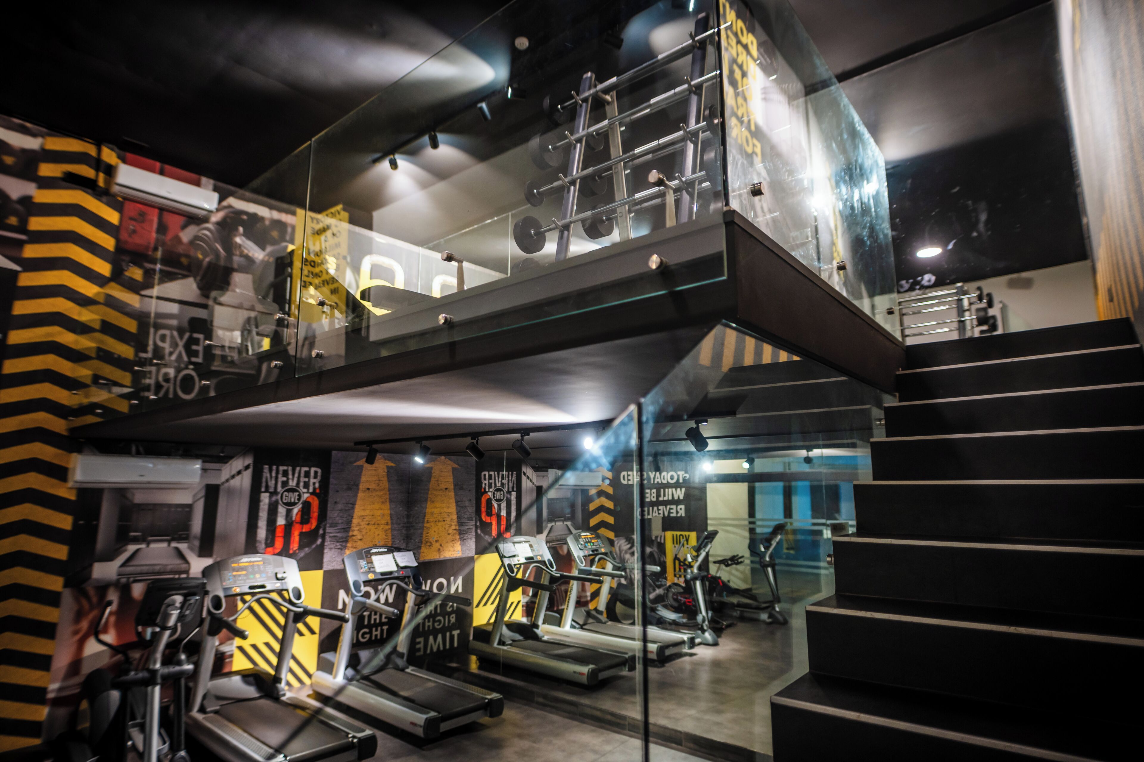 Ein Fitnessstudio mit Laufbändern, Ergometer und Krafttrainingsgeräten. Es gibt zwei Ebenen, die durch eine Glaswand voneinander getrennt sind.