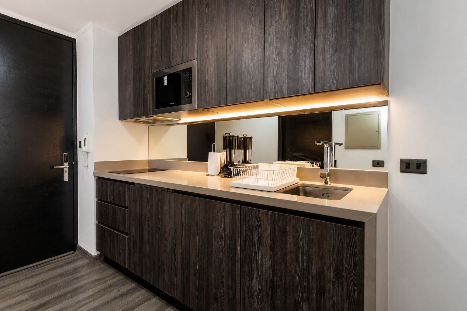 A modern kitchen with dark wood cabinets, a built-in microwave, a stainless steel sink, and a countertop with a dish rack.