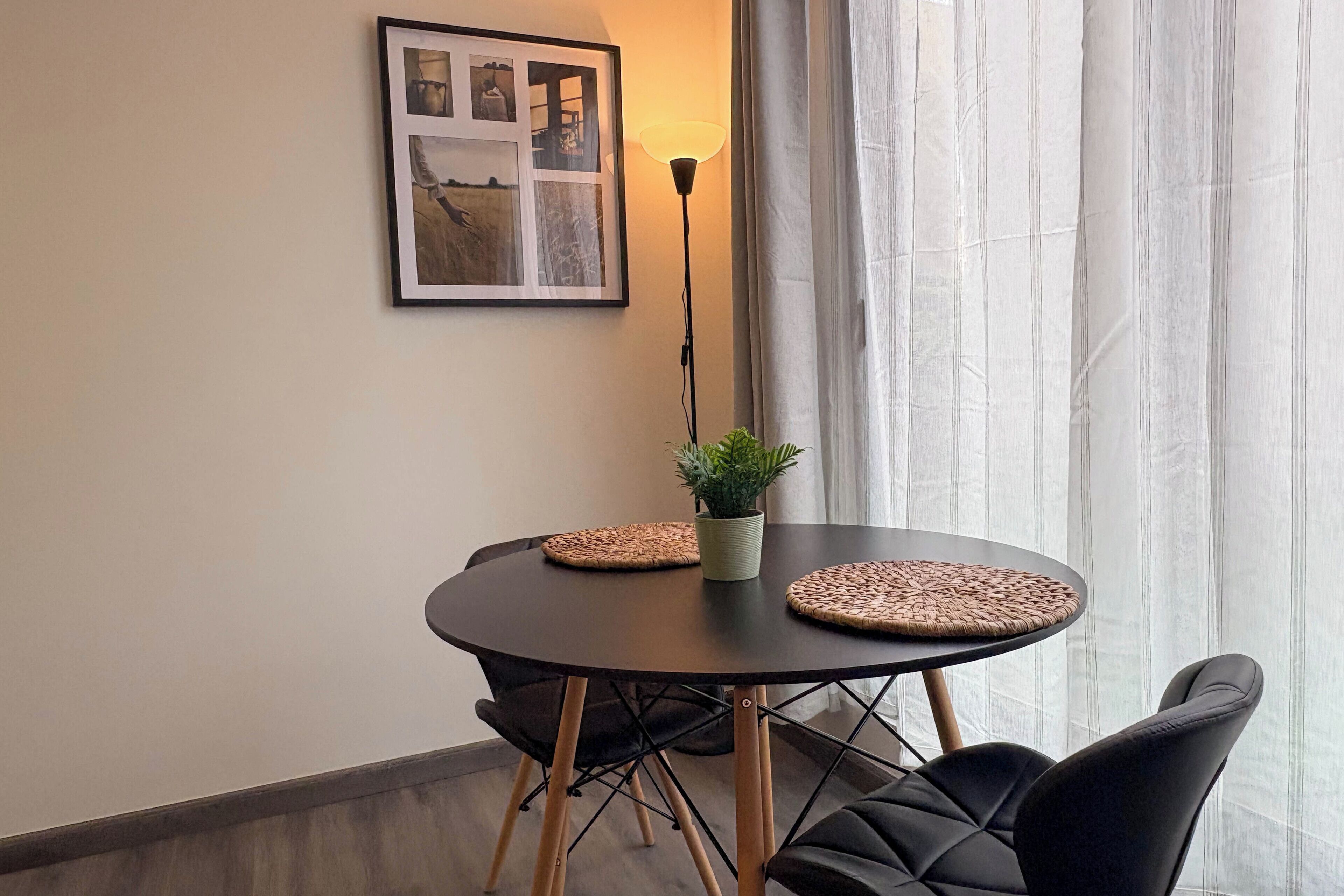 A round table with two woven placemats, a potted plant, and black upholstered chairs. A framed picture hangs on the wall above the table.