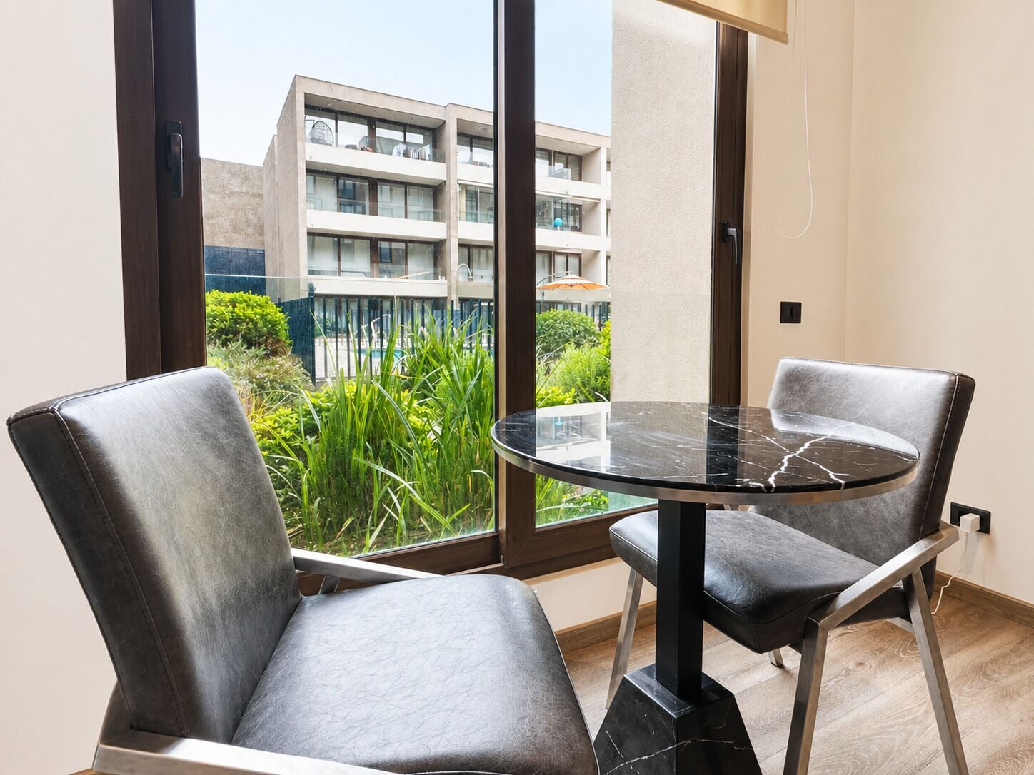 Ein modernes Zimmer mit Glastisch und zwei Ledersesseln, ein großes Fenster mit Blick auf ein Gebäude und Grünflächen.