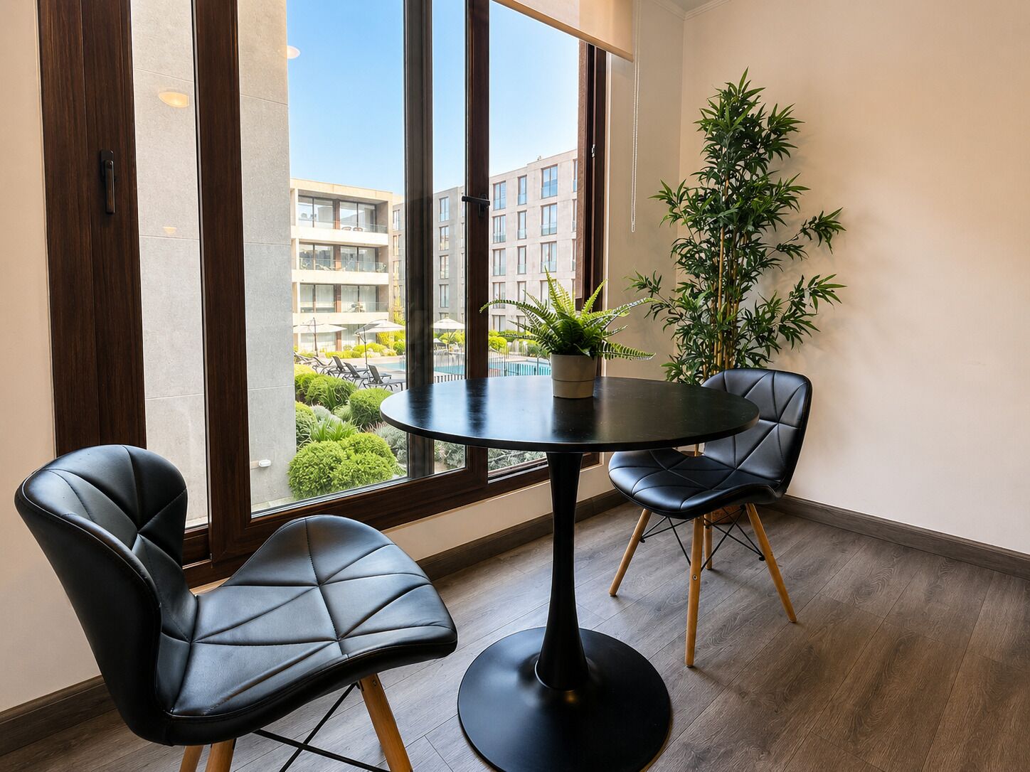 A modern room with a round table, two black leather chairs, a potted plant, and a large window with a view of buildings.