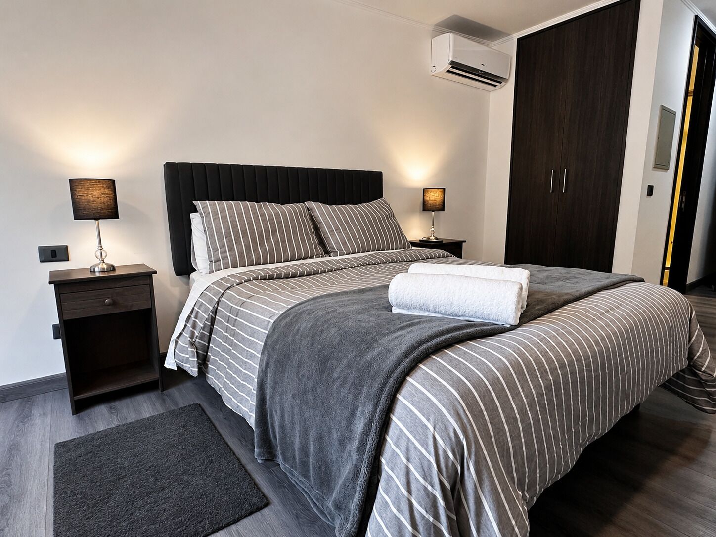 A modern hotel room with a large bed, dark wood wardrobe, bedside lamps, and an air conditioning unit.