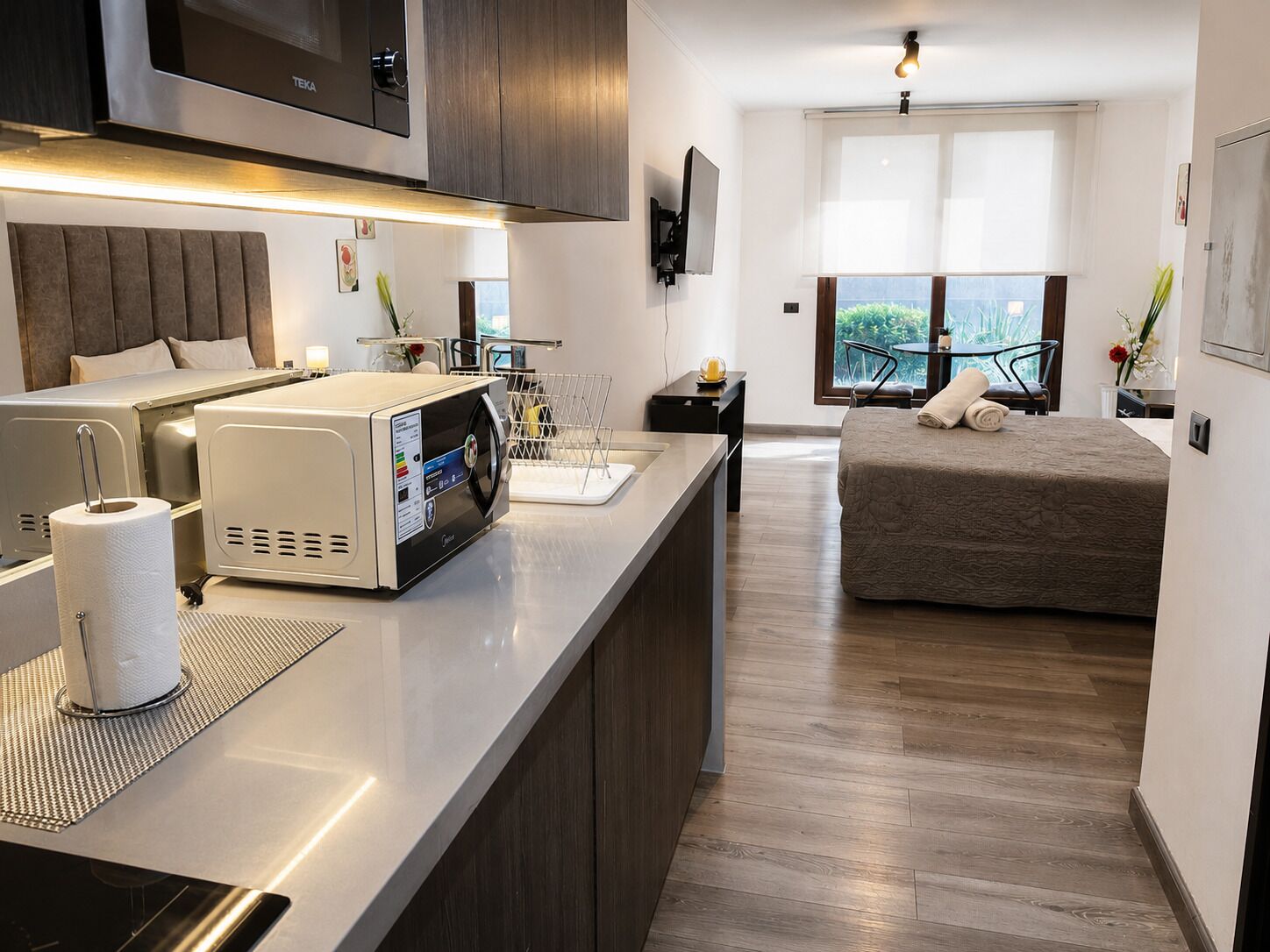 A modern hotel room with a kitchenette, a bed, a TV, and a balcony with a view.