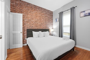 A hotel room with a brick wall, a bed with white linens, a nightstand, a lamp, and a window with curtains. - Walk to Piedmont Park | Free Parking | City Views (Atlanta)