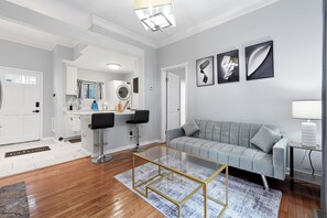 A modern living room with a grey sofa, two black bar stools, a glass coffee table, and a kitchen area with a washing machine. - Walk to Piedmont Park | Free Parking | City Views (Atlanta)