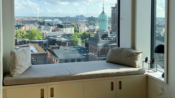 Una habitación con un ventanal grande, un banco empotrado con cojines y vistas a un paisaje urbano.