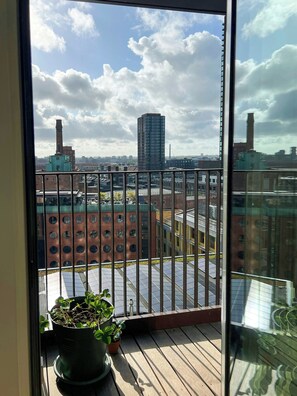 Un balcón con una maceta, vista del paisaje urbano y un edificio con una chimenea alta.