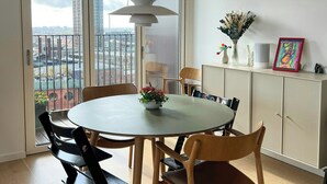 Un comedor con una mesa redonda, sillas, una lámpara colgante, un aparador con adornos, y vistas a edificios a través de una puerta de vidrio.