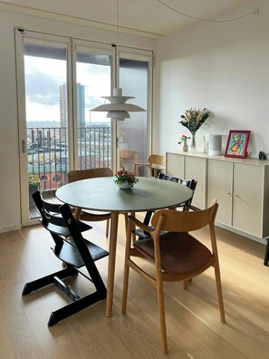En spiseplads med et rundt bord, stole, en hængende lampe, et skab med pynteting og udsigt til bygninger gennem en glasdør.