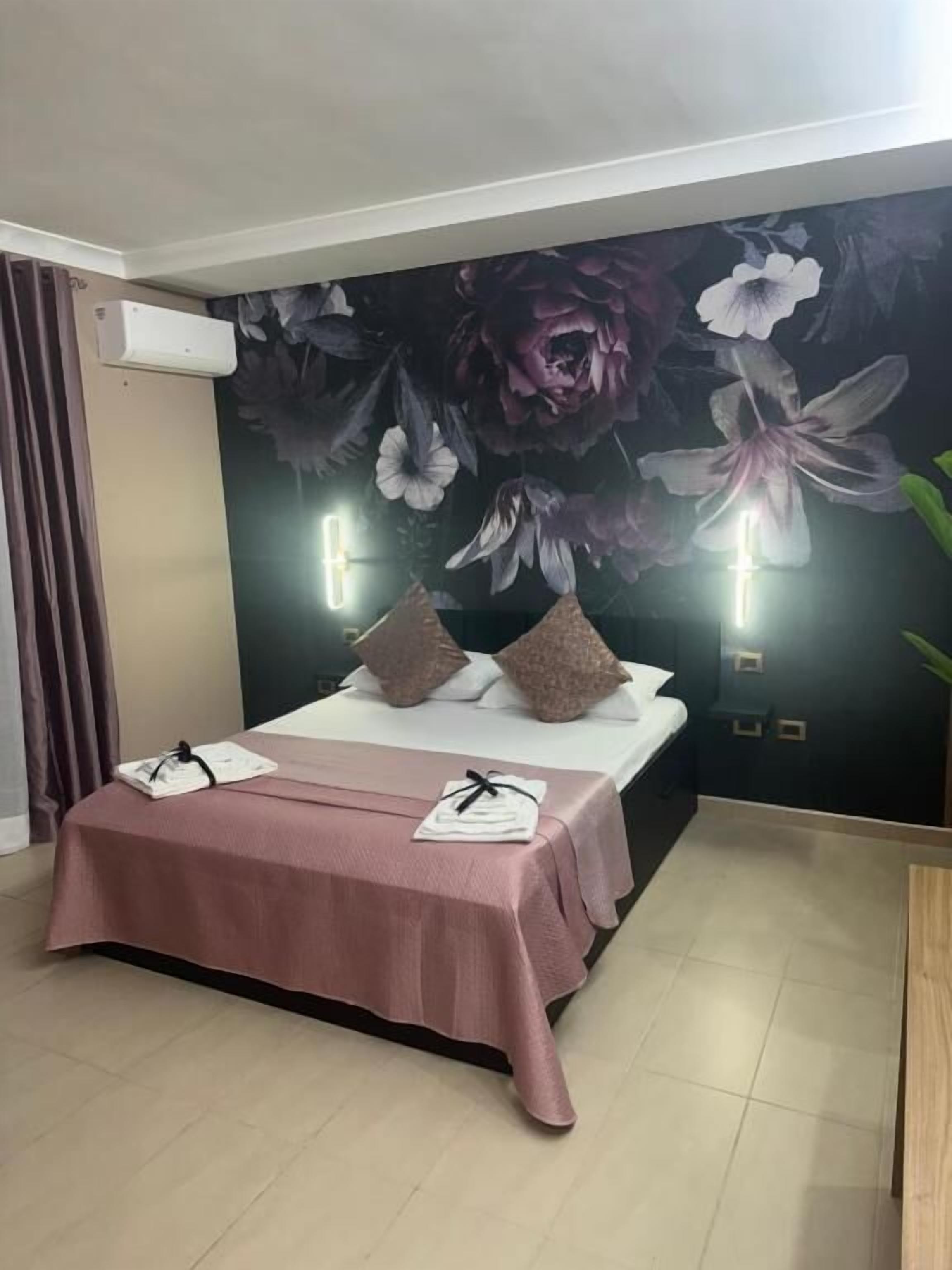 Quarto com cama, almofadas, mesa de cabeceira, ar-condicionado fixado na parede e um mural floral.
