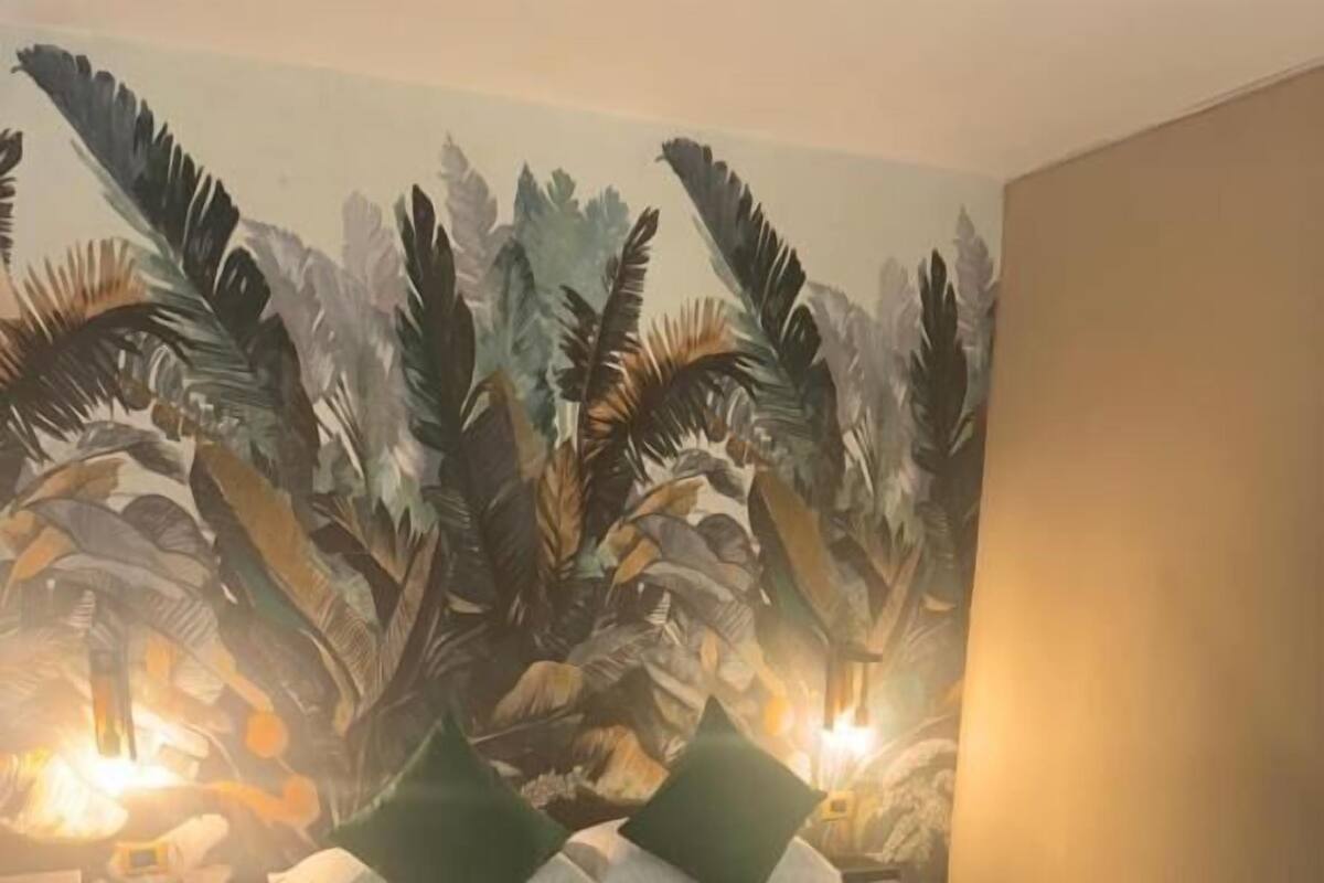 A hotel room with a bed, green pillows, a teal bedspread, and a tropical leaf mural on the wall.