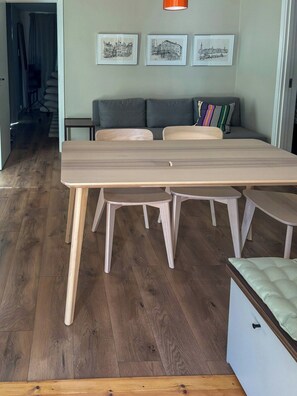 A dining area with a long wooden table, two chairs, a small round table, and a sofa with cushions. - Newly built home with outdoor kitchen on Öland (Mörbylånga)
