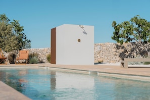 Ein Schwimmbecken mit klarem Wasser, eine Steinmauer und ein weißes Gebäude mit einer Tür und einem kleinen Fenster.