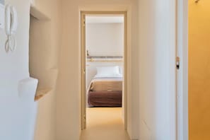 Un estrecho pasillo que lleva a un dormitorio con una cama, un teléfono en la mesita de noche y una estantería.