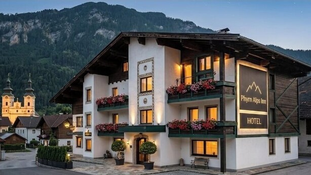 Un hôtel à la façade blanche, doté de balcons ornés de jardinières fleuries, et situé au pied d’une montagne.